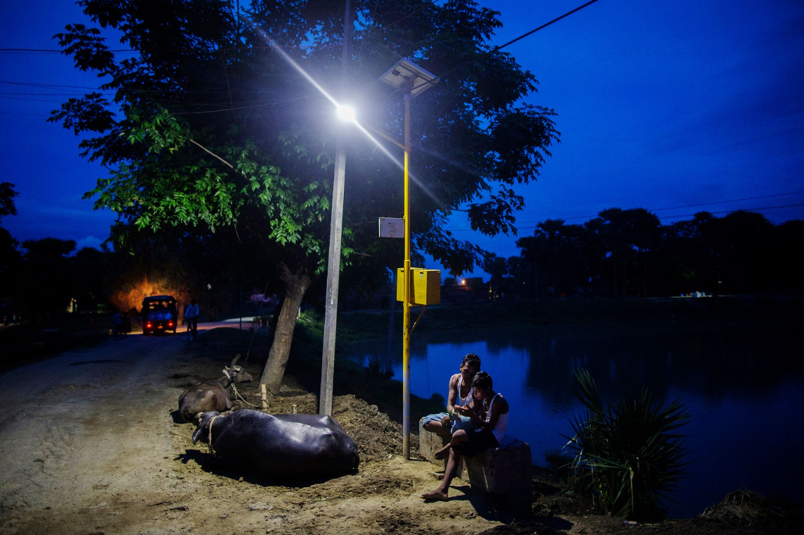 A remote barangay after dark, lit by a single street lamp, where two young people sit at the water's edge looking at a phone